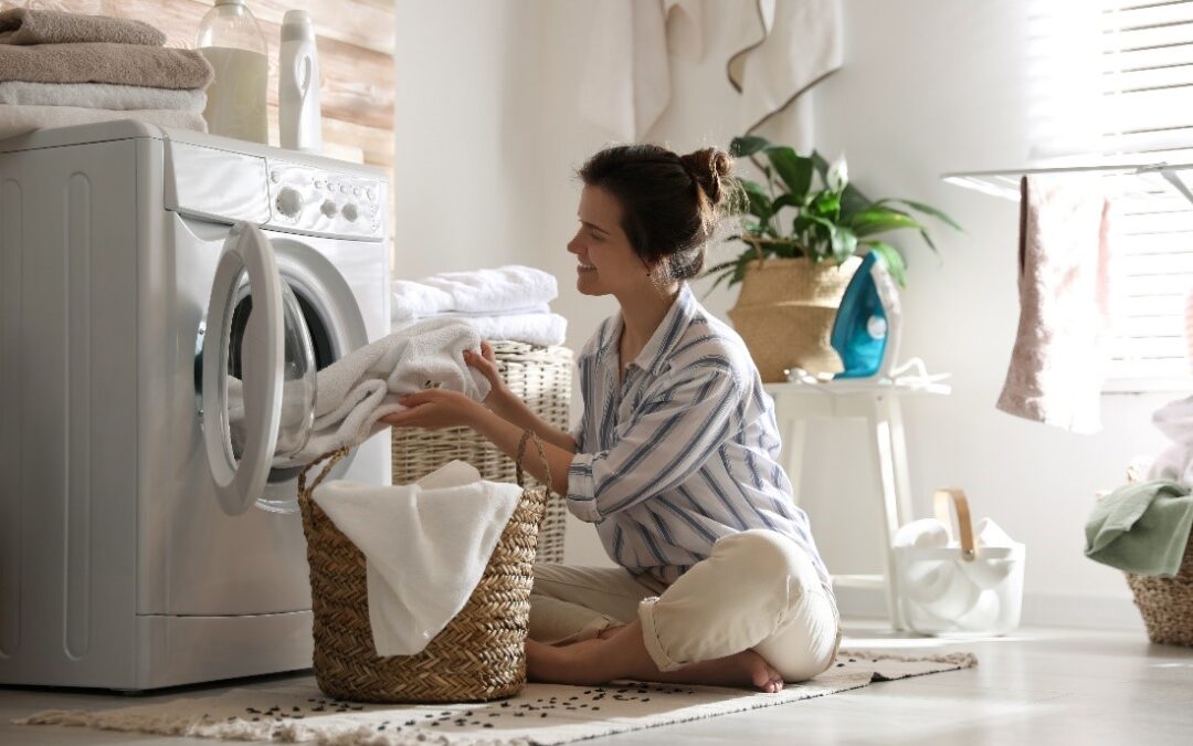 A Smiling Woman in a Striped Shirt Sits on the Floor in Front of a Wicker Laundry Basket and Pulls Towels Out of a Dryer