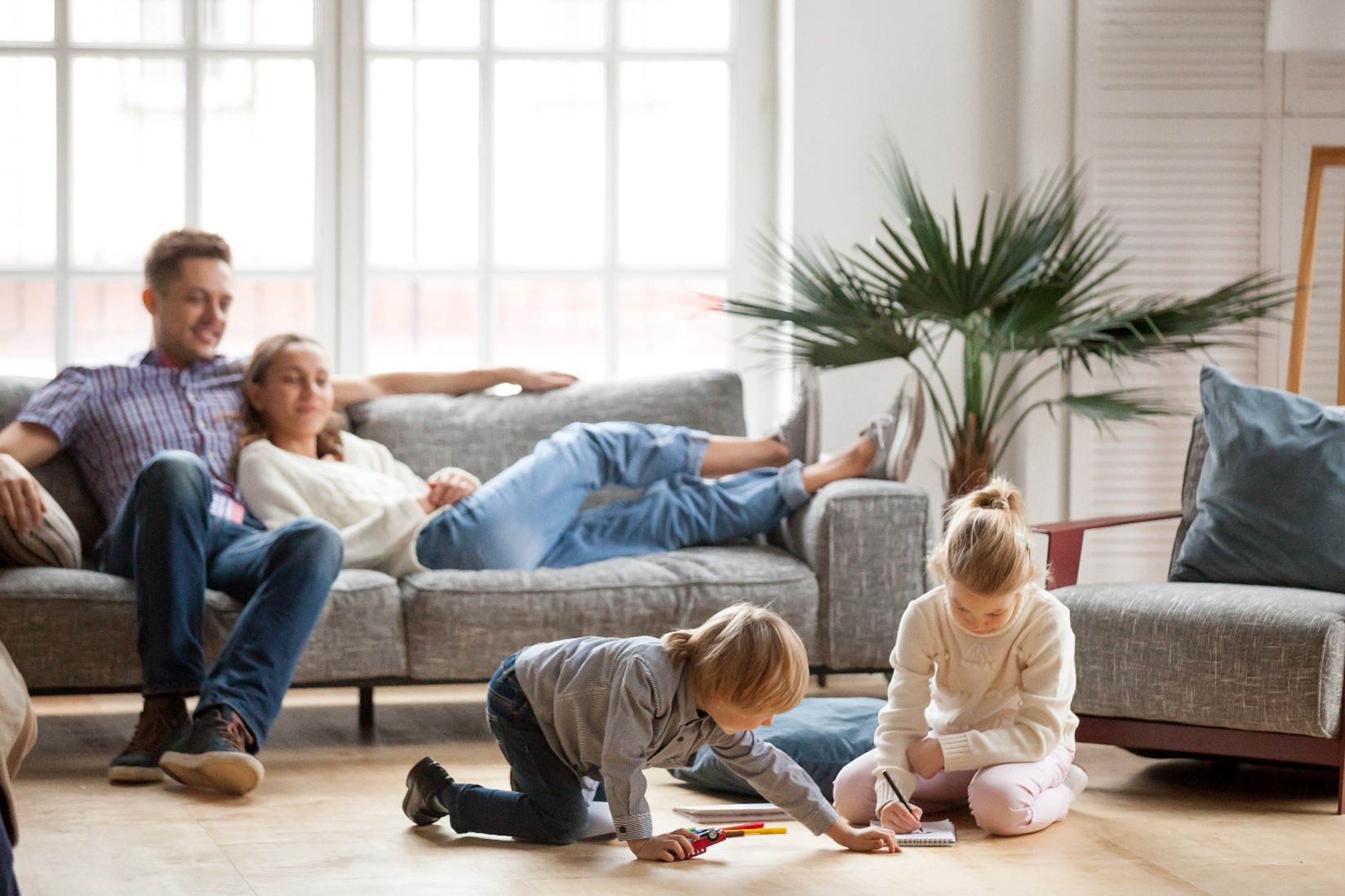 A husband and wife lounging on the couch while their kids sit on the floor while coloring in a healthy indoor environment