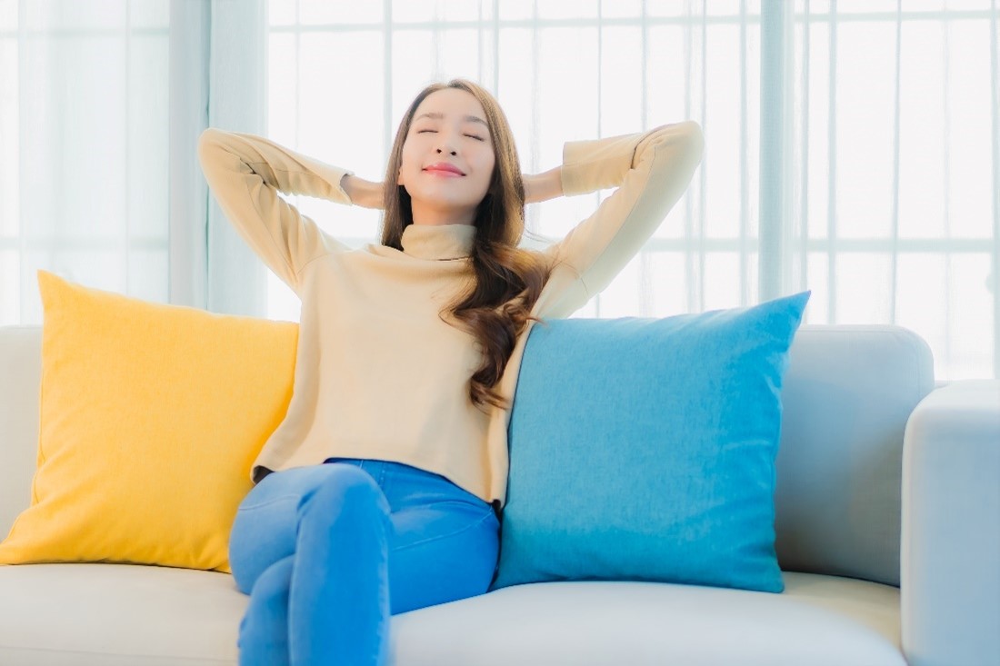 A woman sits on a light gray couch with yellow and blue pillows and reclines with her hands folded behind her head.