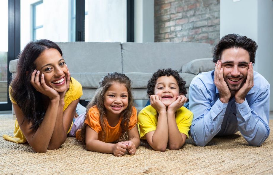 A Dad, Daughter, and Mom Lay on a Beige Carpet Smiling and Embracing Each Other‘s Faces