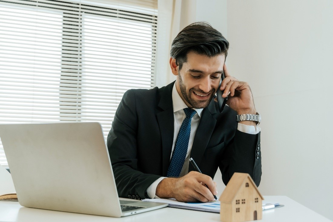 A Man in a Business Suit at a Table with an Open Laptop and a House Figurine on it While on the Phone and Taking Notes