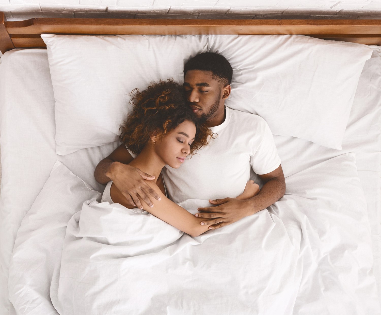 A Young Adult Couple Peacefully Sleeping in an Embrace in White Bedding with a Wooden Headboard Behind Them