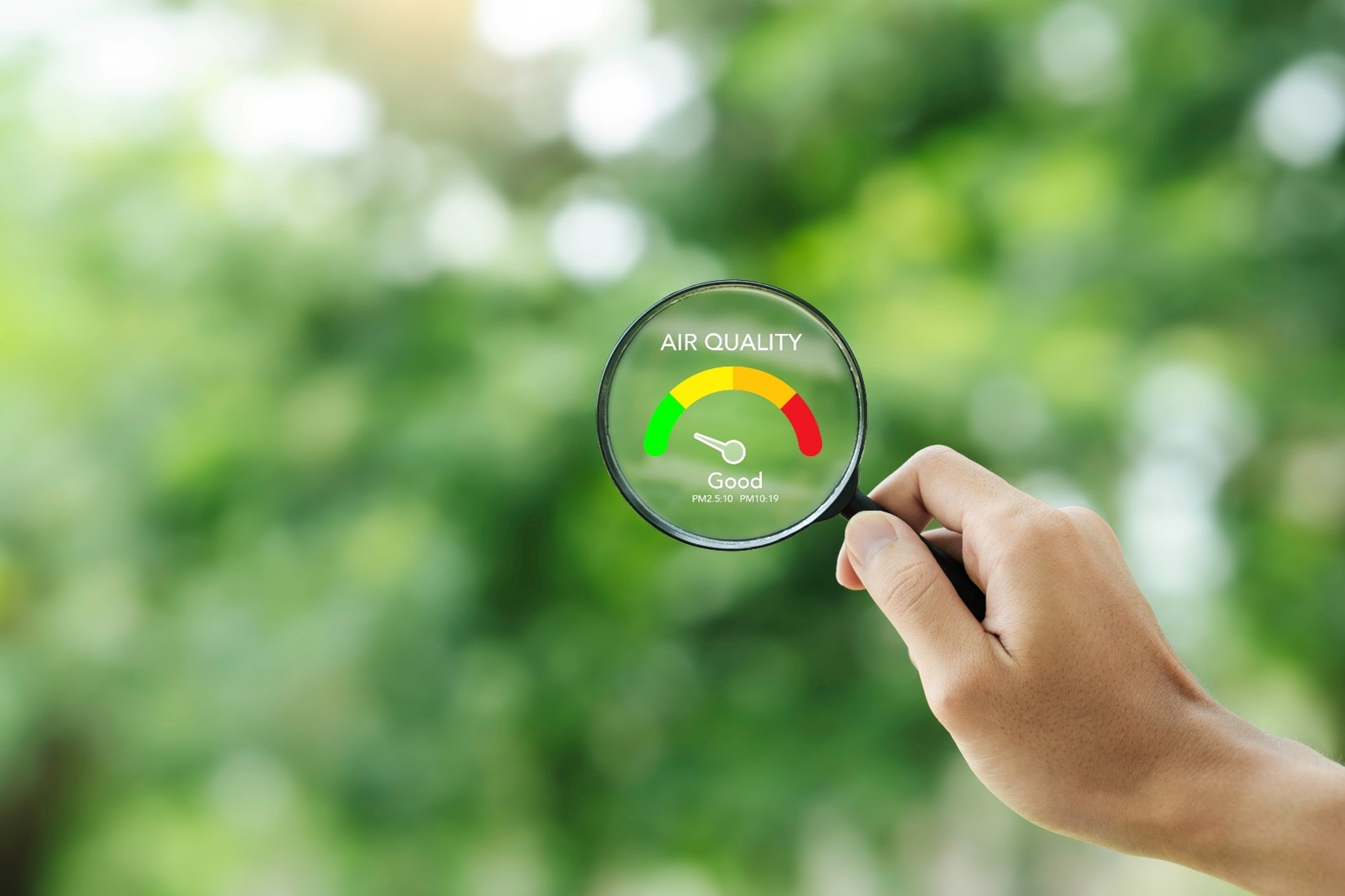 A hand holding a magnifying glass showing good air quality next to a blurred background of greenery.